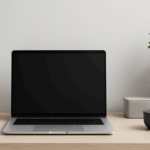 北欧風のシンプルで整然としたデスク上に、黒い画面のノートパソコン、黒いテーブルランプ、白いマグカップ、グレーの収納ボックス、白い鉢植えに入った観葉植物がバランスよく配置されている、モノトーン基調のモダンなワークスペース。