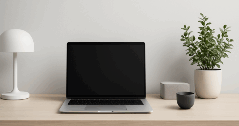 北欧風のシンプルで整然としたデスク上に、黒い画面のノートパソコン、黒いテーブルランプ、白いマグカップ、グレーの収納ボックス、白い鉢植えに入った観葉植物がバランスよく配置されている、モノトーン基調のモダンなワークスペース。