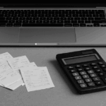 整然としたデスク上に、ノートパソコン、電卓、領収書、ノート、会計帳簿がモノクロで配置され、右奥には観葉植物が置かれている。経理業務の雰囲気を表すプロフェッショナルでミニマルな構成。