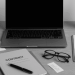 デスクの上にノートパソコン、契約書と記載された書類、クリップボード、メガネ、観葉植物が整然と配置されたモノクロ写真。契約書作成やチェックを想起させる、プロフェッショナルで信頼感のある構図。