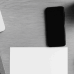 左上にノートパソコンの一部が写り、右側にはスマートフォン、白いマウス、観葉植物が配置されたモノクロのデスク風景。中央は空白の用紙とノートが整然と置かれ、営業活動やタスク管理を連想させるシンプルな構図。