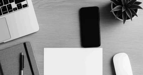 左上にノートパソコンの一部が写り、右側にはスマートフォン、白いマウス、観葉植物が配置されたモノクロのデスク風景。中央は空白の用紙とノートが整然と置かれ、営業活動やタスク管理を連想させるシンプルな構図。