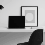 北欧風のミニマルなワークスペース。白いデスクに黒いノートパソコン、モダンなデスクライト、抽象画のアートが配置され、整然とした雰囲気が漂う。