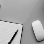 ライトグレーのデスクにノートパソコン、開かれたノートと黒いペン、白いワイヤレスマウス、スマートフォン、観葉植物が整然と配置されたモノクロ写真。事務処理や日常業務の落ち着いた雰囲気を演出するシンプルな構成。