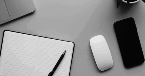 ライトグレーのデスクにノートパソコン、開かれたノートと黒いペン、白いワイヤレスマウス、スマートフォン、観葉植物が整然と配置されたモノクロ写真。事務処理や日常業務の落ち着いた雰囲気を演出するシンプルな構成。